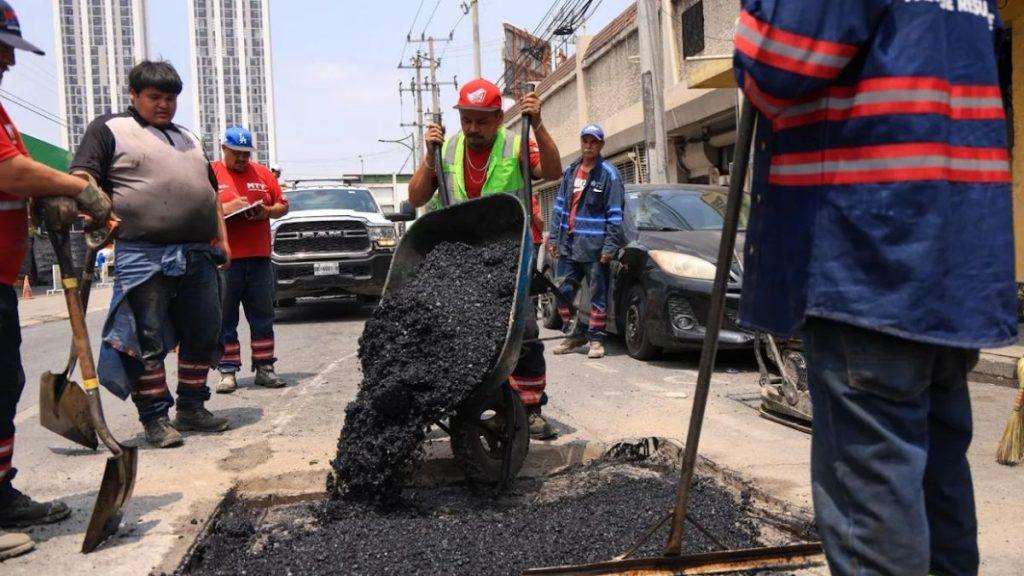Road construction crew pours hot asphalt from a wheelbarrow into a street hole while others supervise in an urban area with vehicles parked nearby and tall buildings in the background   Informe Regio