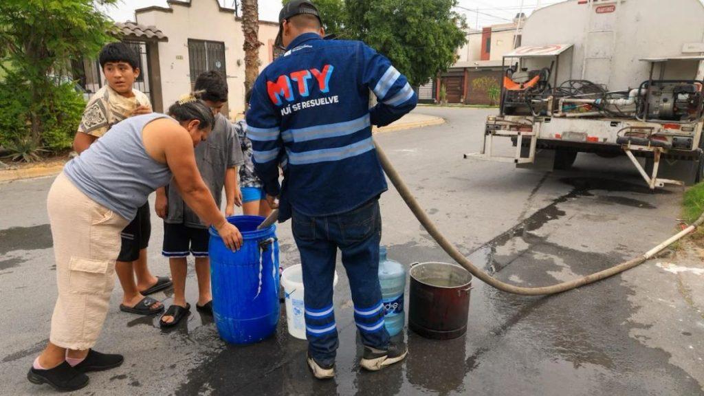 Emergency responder in blue uniform distributes water to a blue barrel as a family watches on a street A tanker truck with a hose is in the background   Informe Regio