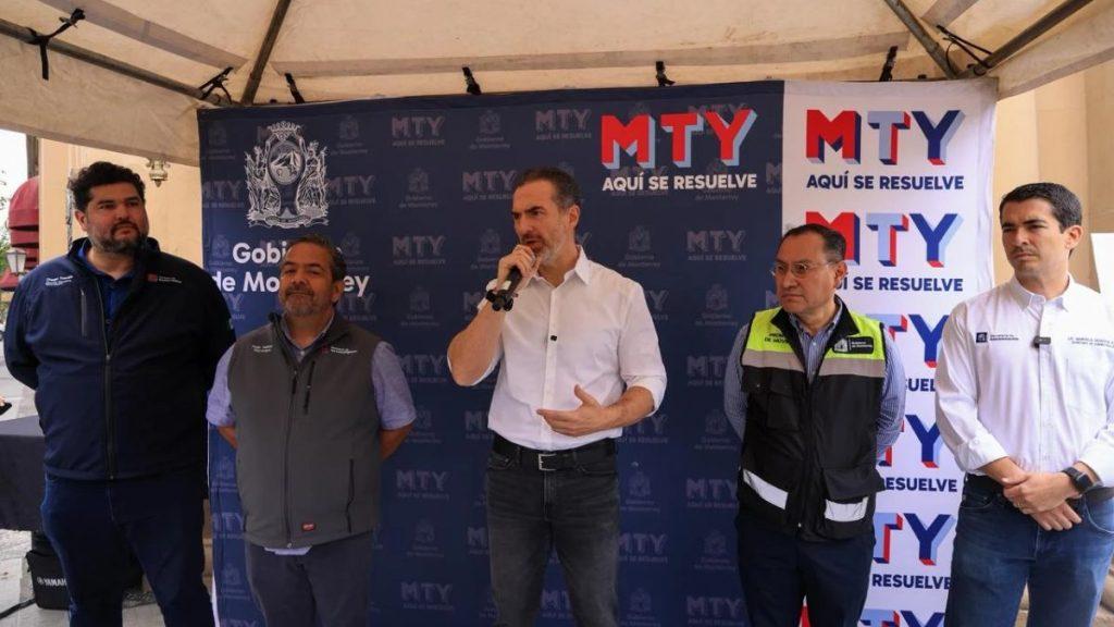 Five men stand on a stage under a tent with a blue MTY backdrop center figure in a white shirt speaks into a microphone while others listen nearby   Informe Regio