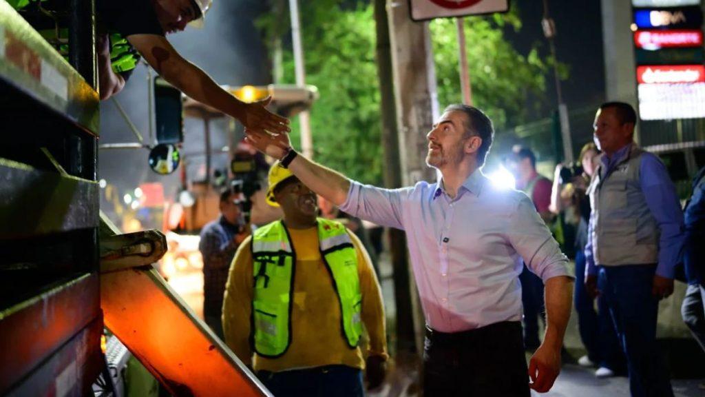 Man in a pale shirt shakes hands with a construction worker wearing a neon vest beside heavy equipment at night   Informe Regio