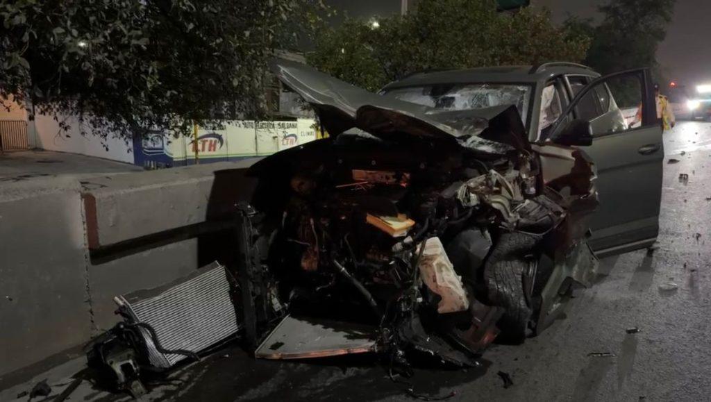 Nighttime car crash severely damaged gray sedan with front end destruction deployed airbags and exposed engine debris on wet road and a concrete barrier to the left emergency lights in the distance   Informe Regio