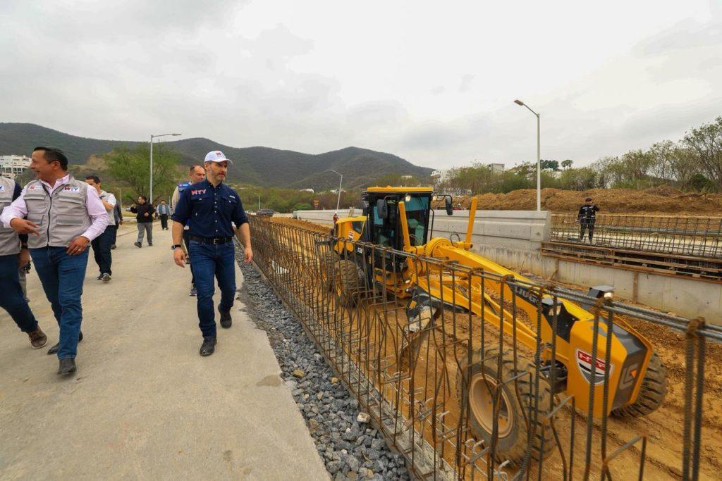 Group of construction workers walking along a concrete path beside a large yellow excavator and exposed rebar at a trench site with hills in the background   Informe Regio