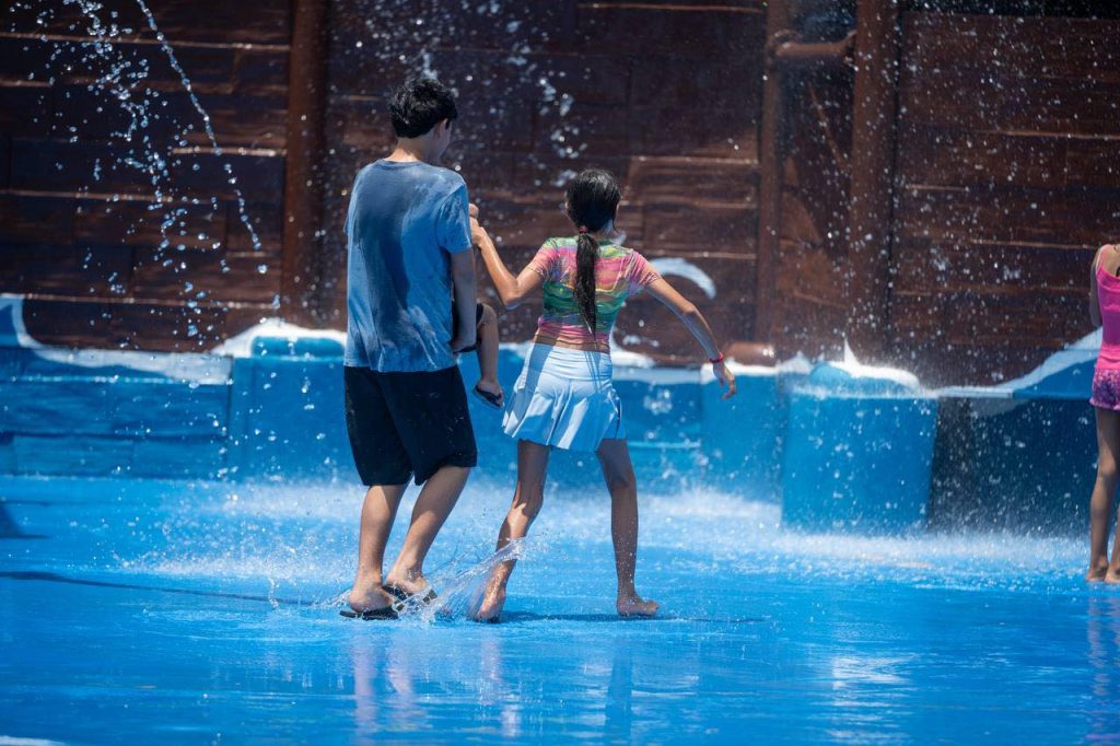Two children walk hand in hand through a shallow splash pad water spraying around them in a sunny play area   Informe Regio