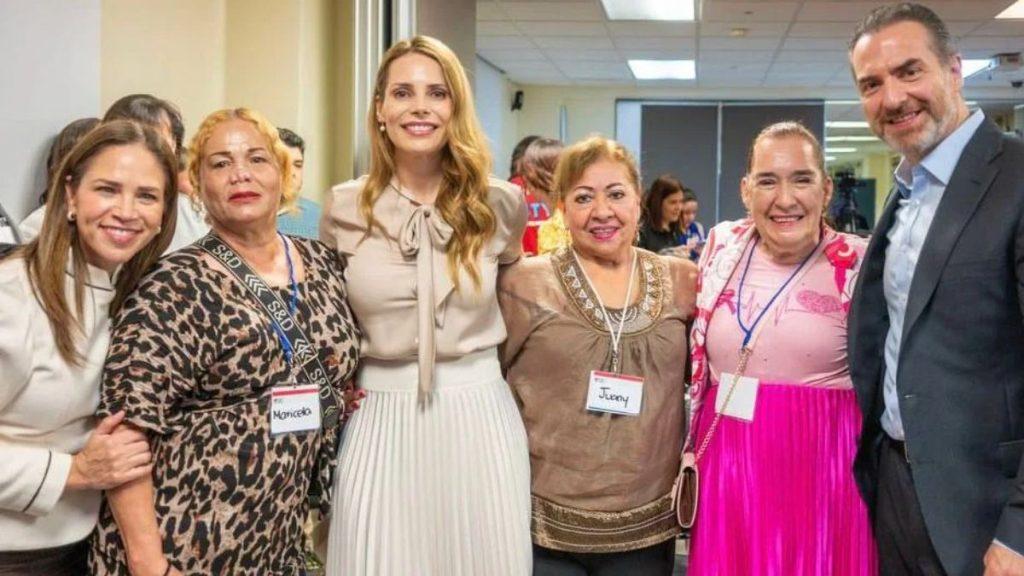 Six adults pose for a group photo in a hallway smiling at the camera at a social event central tall blonde woman in beige blouse and pleated skirt stands with five others wearing name tags   Informe Regio
