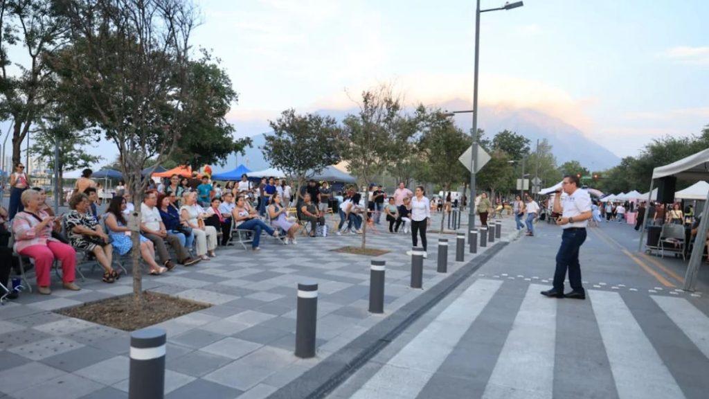 Man with a microphone speaks to a seated crowd at an outdoor event on a paved promenade with vendor tents in the background under trees   Informe Regio