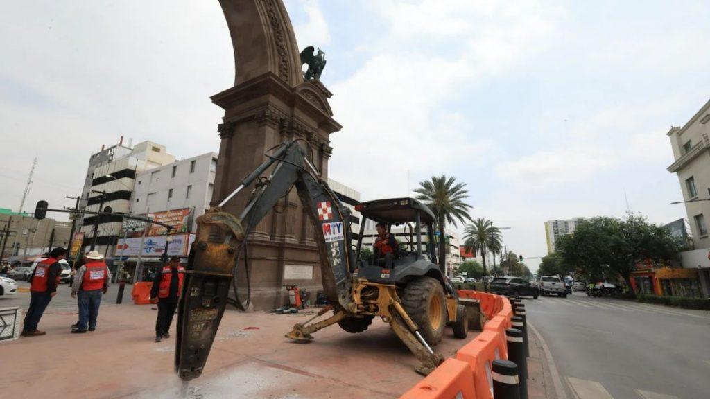 Construction work around a tall stone arch monument on a city street with a backhoe loader and workers in red vests nearby   Informe Regio