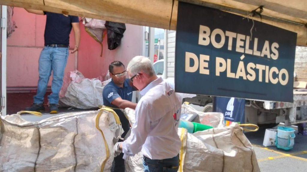 Two men unloading large white bags of plastic bottles from a truck under a sign that reads Botellas de plástico   Informe Regio