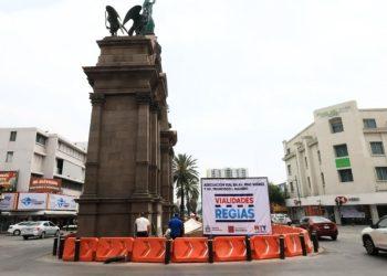 Tall stone monument rising from a traffic circle surrounded by bright orange barrier blocks and a large billboard that reads Vialidades Régias   Informe Regio