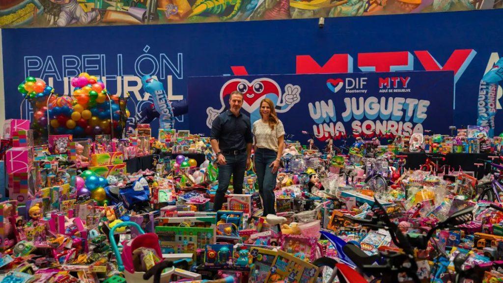 Man and woman pose amid a mountain of donated toys at a charity event banner reads Un Juguete Una Sonrisa in the background   Informe Regio