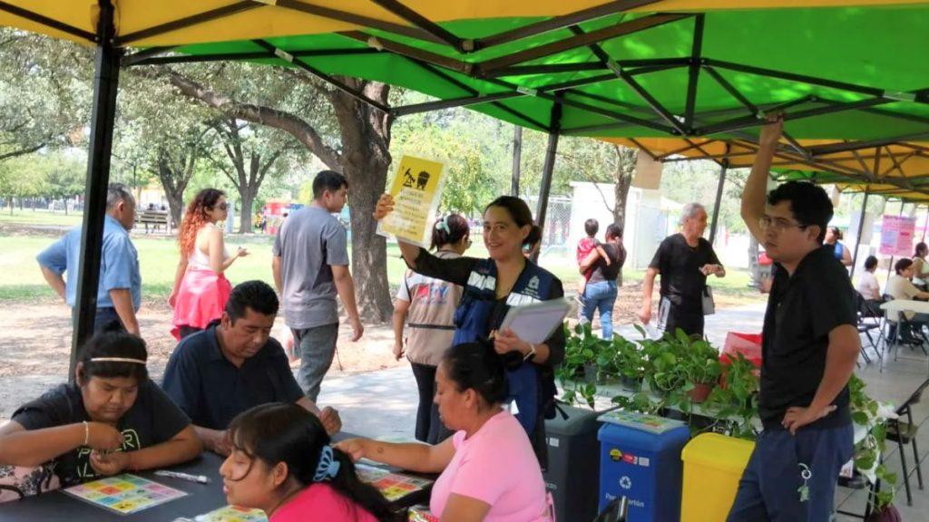 People at an outdoor community fair under colorful tents a woman raises a flyer while others sit at a craft table in a park setting   Informe Regio