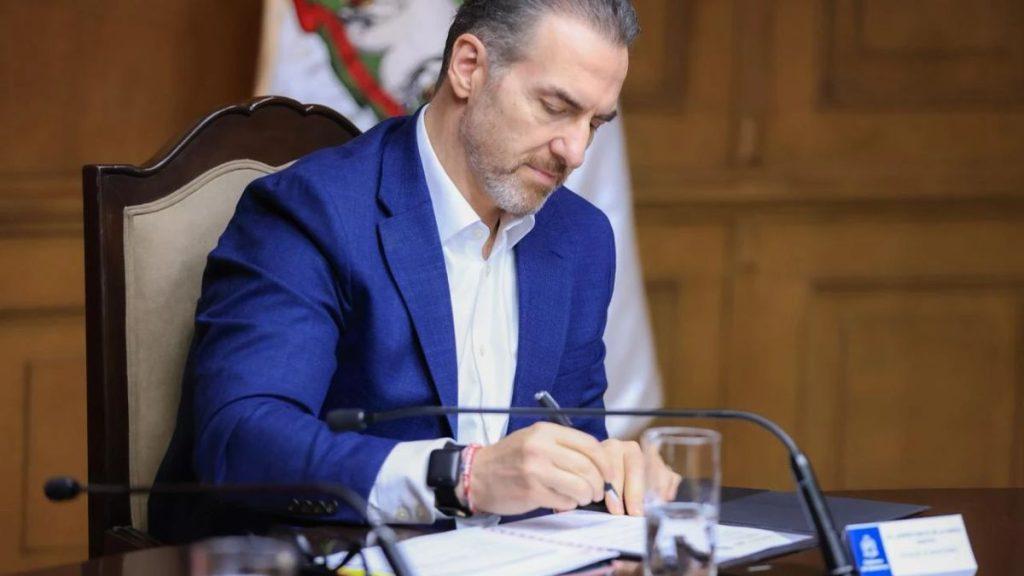 Adrián-de-la-garza-convenio-inea-educación-básica-primaria-secundaria-monterrey - Informe Regio Man in a blue suit signs documents at a desk in a formal meeting room with a flag in the background Informe Regio