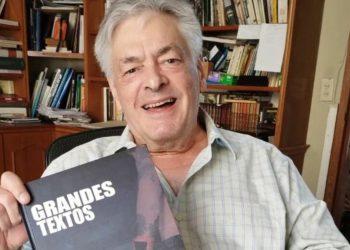 Older man with gray hair smiles at the camera while holding a book titled GRANDES TEXTOS   Informe Regio