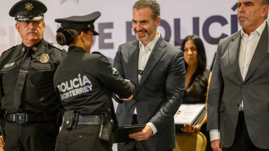 Secretaría-de-seguridad-y-protección-ciudadana-adrián-de-la-garza - Informe Regio Man in a gray suit shakes hands with a Monterrey police officer in uniform during a formal ceremony with other officials nearby on stage Informe Regio