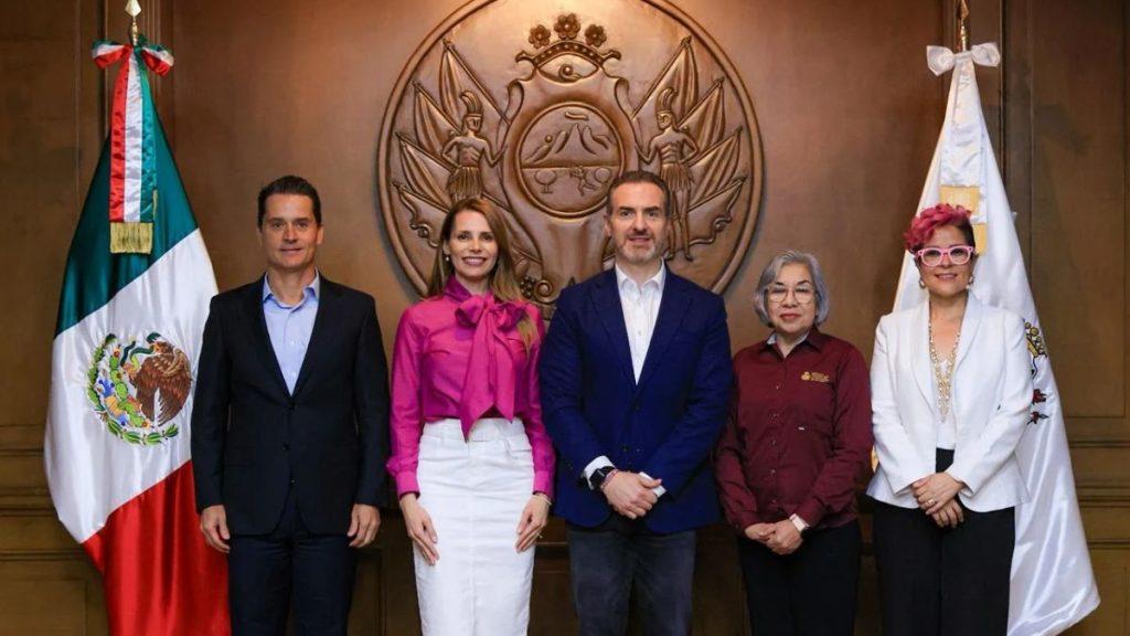 Adrián-de-la-garza-convenio-gaby-oyervides-inea - Informe Regio Five people in formal attire pose for an official group photo in a government chamber with a Mexican flag and a large seal backdrop behind them Informe Regio