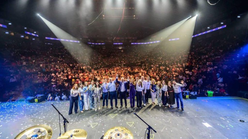 Group of performers and staff posing on a large stage as a big audience cheers in a packed arena at a concert or event   Informe Regio