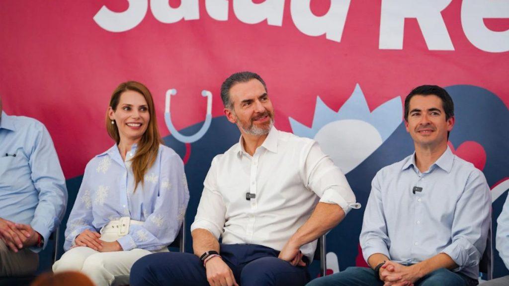 Four panelists sit on a stage in front of a pink backdrop engaged in a discussion center man smiles woman in blue blouse to his left man in light blue shirt to his right   Informe Regio