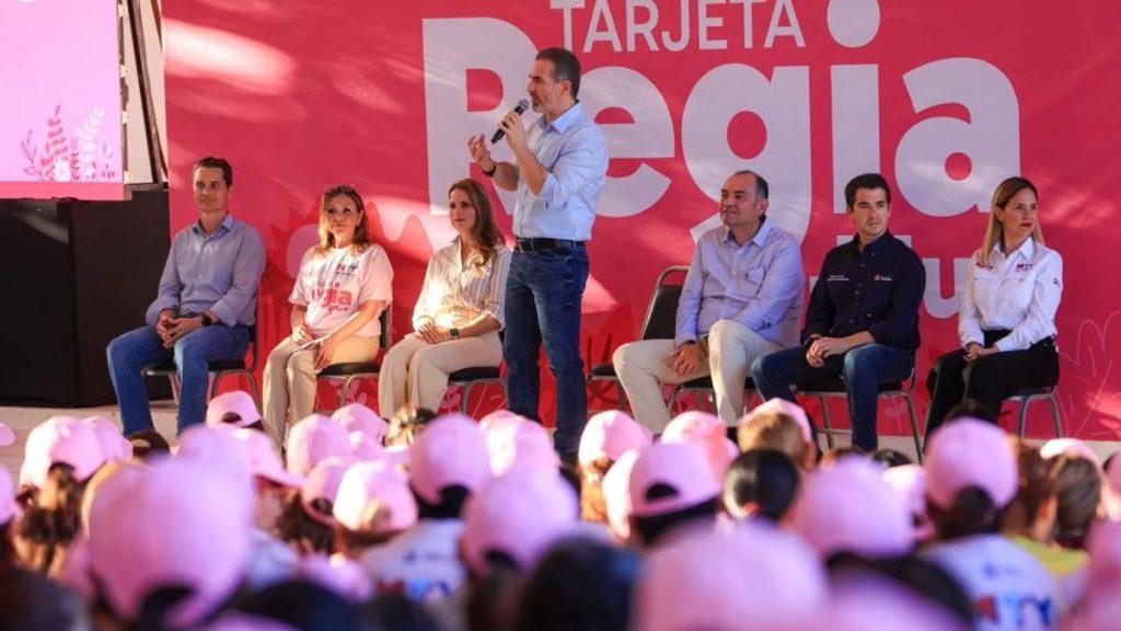 Panel of six people seated on a stage as a man speaks into a microphone audience wearing pink hats in front of a pink backdrop   Informe Regio