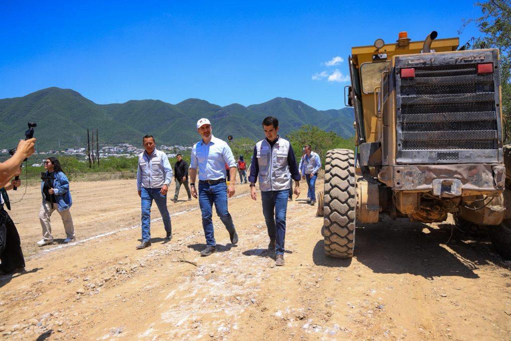 Group of people walking on a dusty construction site with a large yellow dump truck to the right and green mountains in the background   Informe Regio