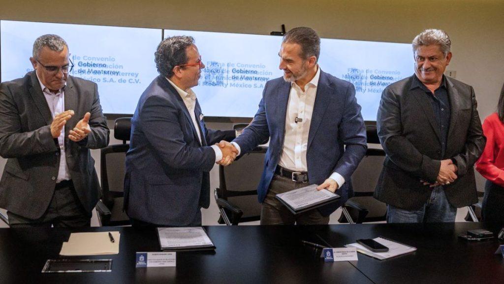Two men in business suits shake hands across a table after signing documents others applaud in the background   Informe Regio