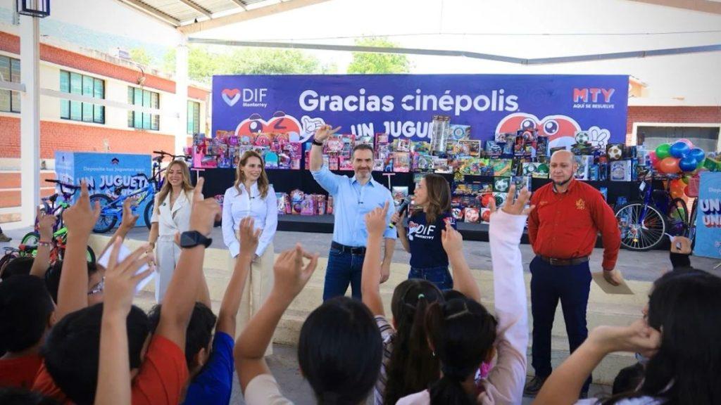 Group of adults on stage at a charity event with a blue banner reading Gracias Cinépolis kids in foreground raising hands enthusiastically introduced by a presenter wearing a light blue shirt   Informe Regio