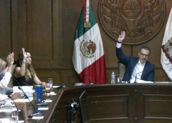 A formal government meeting with a man in a suit raising his hand at a long wooden dais Mexican flag in the background   Informe Regio