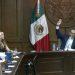 A formal government meeting with a man in a suit raising his hand at a long wooden dais Mexican flag in the background   Informe Regio