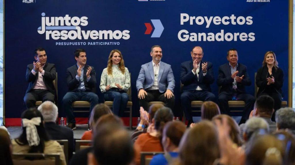 El-alcalde-adrian-de-la-garza-y-su-trabajo-en-monterrey - Informe Regio Panel of seven speakers on a stage applauding at a conference with a dark blue backdrop that reads juntos resolvemos and Proyectos Ganadores Informe Regio