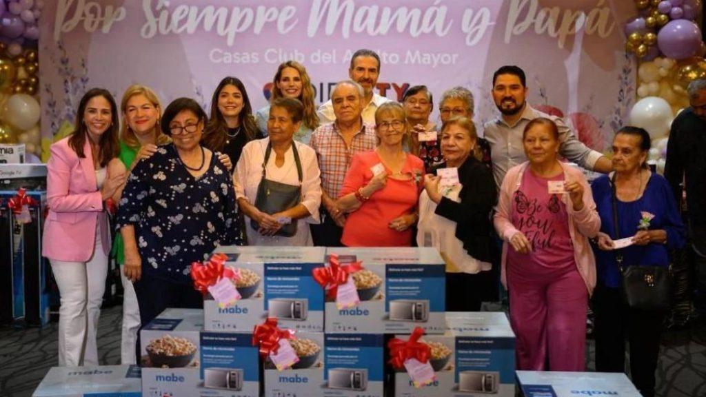 Group of people posing for a photo at a charity event with stacked gift boxes wrapped in red ribbons in front of a pink banner and balloons   Informe Regio