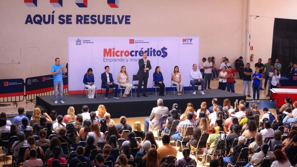 Stage event about microcréditos a speaker stands while others sit on chairs banner reads Microcrédito$ Emprende y resuelve with an audience watching   Informe Regio