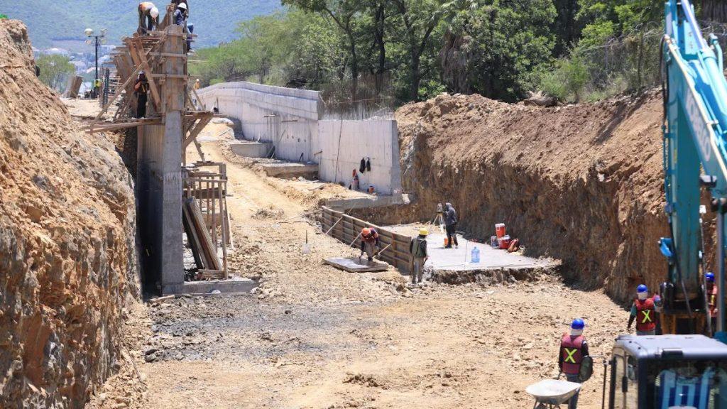 Obras-circuito-vial-huajuco-av-acueducto - Informe Regio Construction site with workers in safety vests building a concrete retaining wall along a trench scaffolding and heavy machinery visible on both sides Informe Regio