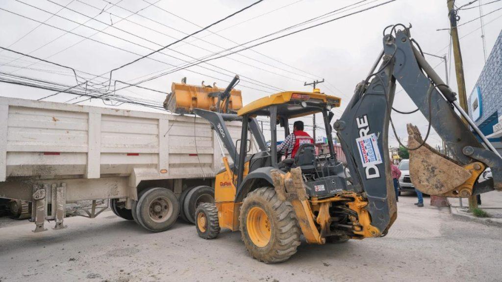 Yellow backhoe loader scooping soil into a large dump truck on a dusty street with overhead power lines and a worker in a red vest nearby   Informe Regio