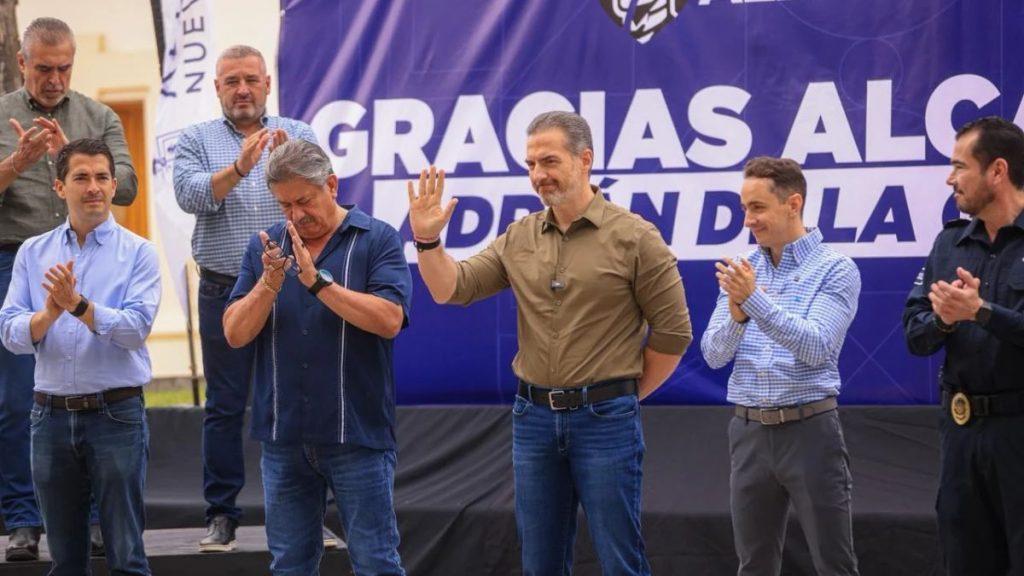 Group of men on a stage at a public event the central man waves while others clap with a blue banner behind reading GRACIAS   Informe Regio