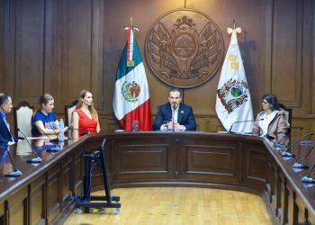A group of officials seated around a large wooden conference table in a formal government chamber with the Mexican flag behind the speaker   Informe Regio