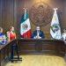 A group of officials seated around a large wooden conference table in a formal government chamber with the Mexican flag behind the speaker   Informe Regio