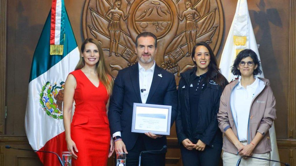 Four people pose with a framed certificate in a formal room with a Mexican flag behind them and a carved emblem on the wall   Informe Regio