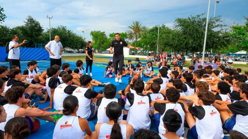 Apertura-de-nueva-clinica-de-basquetbol - Informe Regio Coach speaks to a large group of young athletes seated on a blue outdoor court for a training session Informe Regio