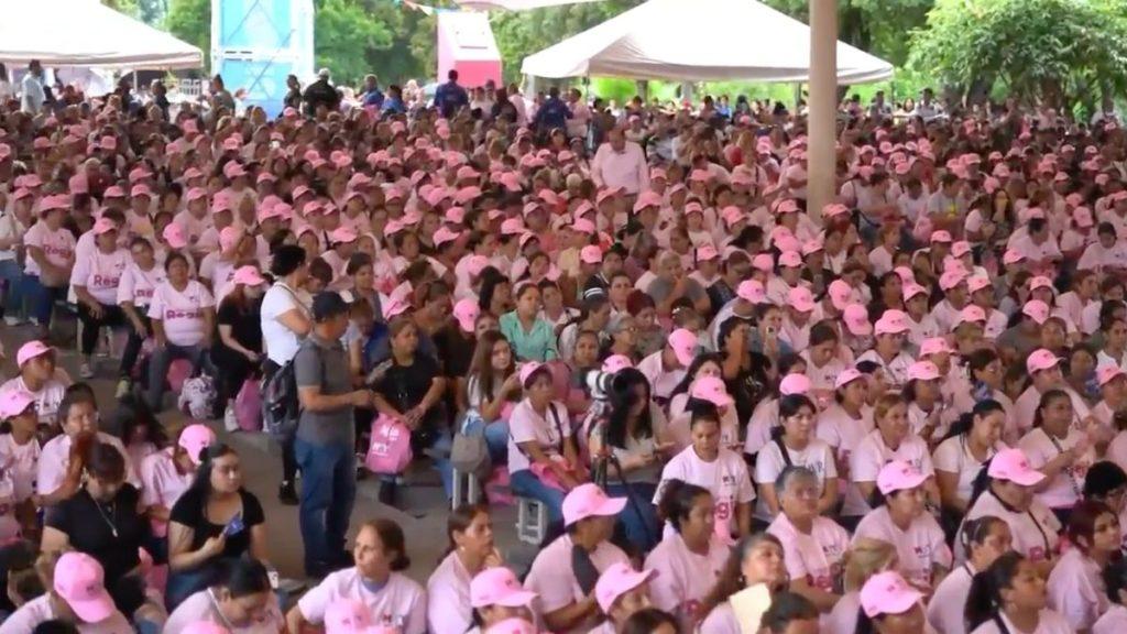 Apoyo-economico-para-mujeres - Informe Regio Large crowd of people in pink shirts and pink caps seated outdoors under tents at an event park Informe Regio