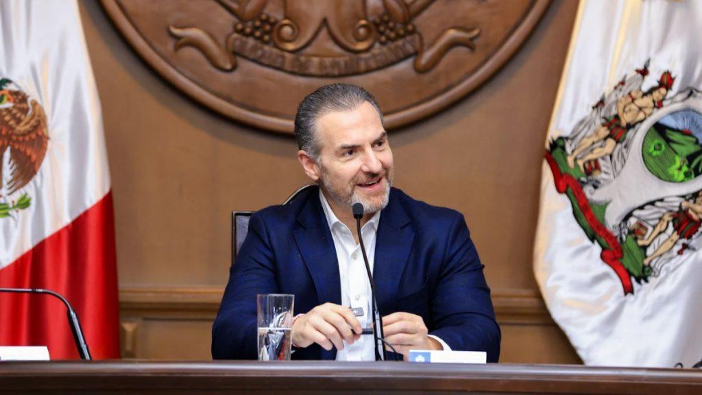 Man in a navy suit speaks into a microphone at a formal government meeting with flags and a crest behind him   Informe Regio
