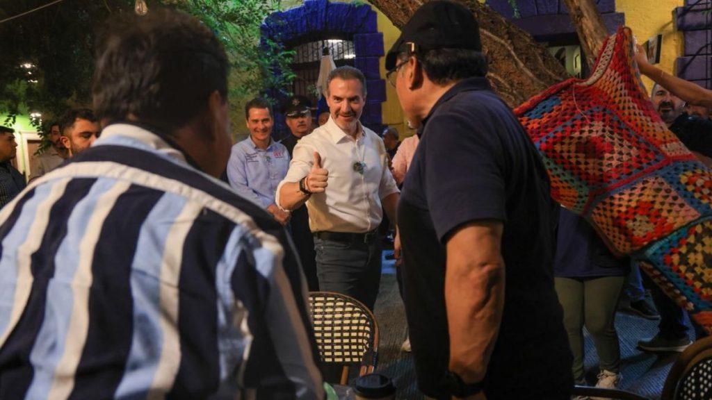 Man in white shirt giving a thumbs up in a lively outdoor gathering surrounded by people and a colorful textile backdrop   Informe Regio