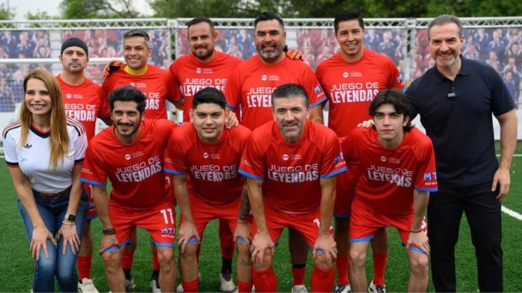 Group of players in red Juego de Leyendas soccer jerseys pose for a team photo on a green field with a woman in white on the left and a man in black on the right   Informe Regio