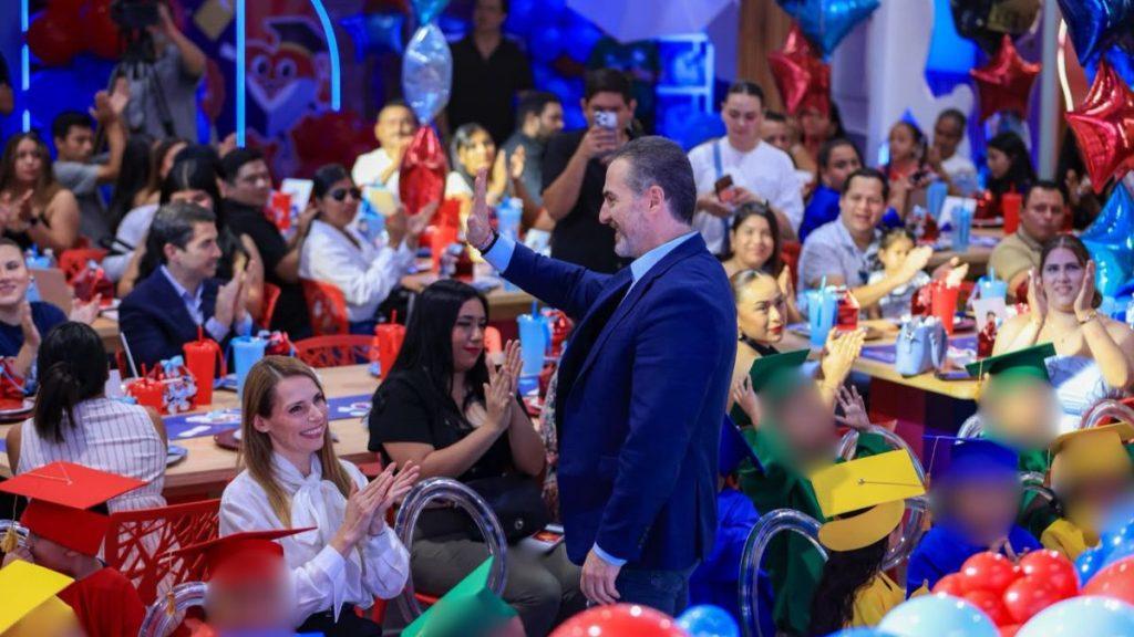 Man in a blue suit waves to a cheering crowd at a festive indoor event with colorful balloons and graduates in caps nearby   Informe Regio