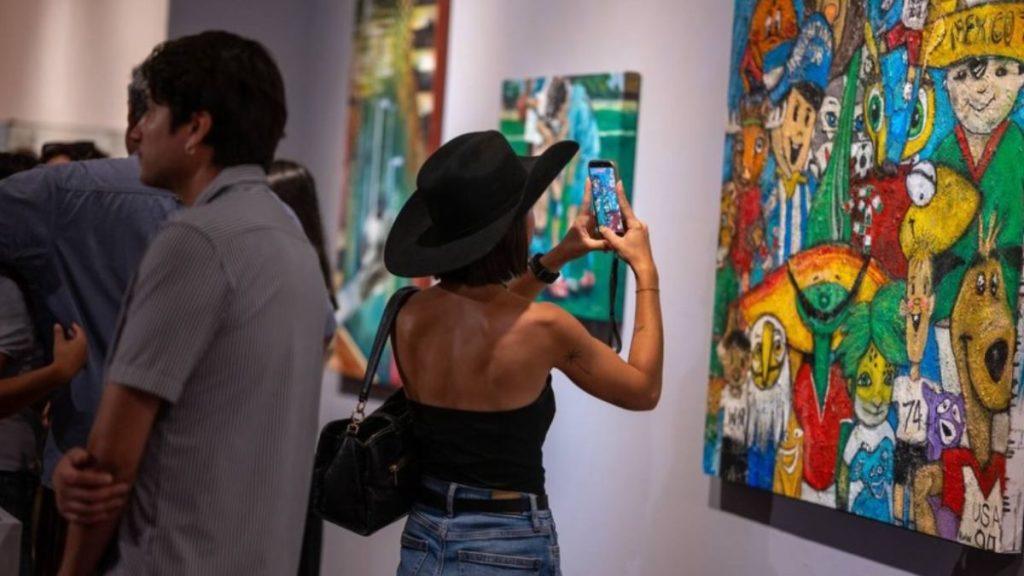 Woman in a black wide brimmed hat takes a photo of a colorful mural with her phone in an art gallery   Informe Regio