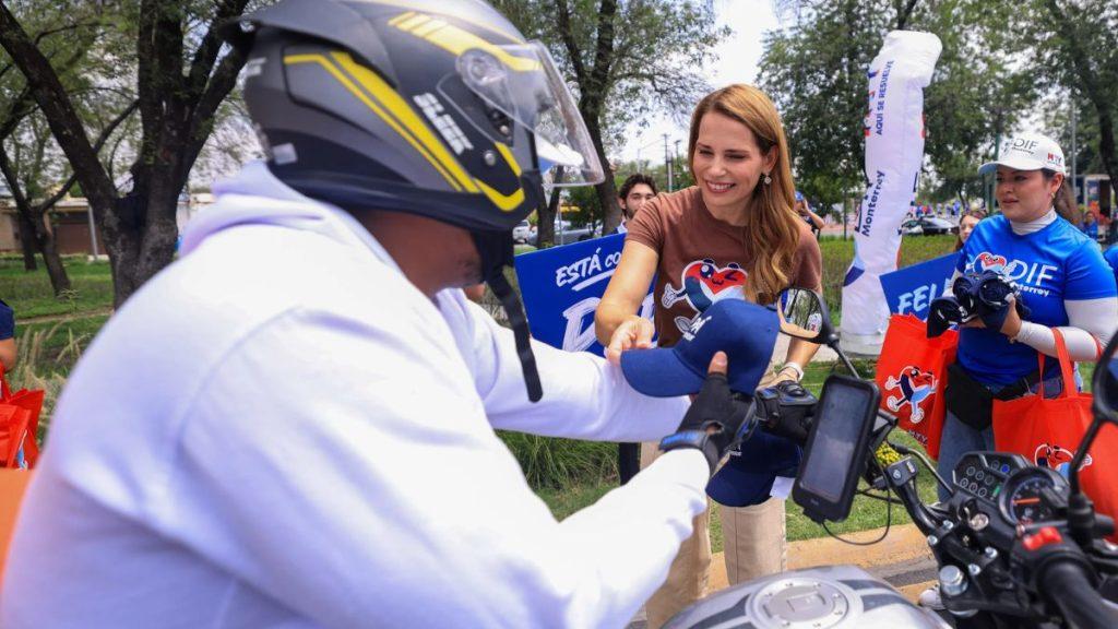 Gaby-oyervides - Informe Regio A woman in a brown shirt hands a blue cap to a motorcyclist wearing a helmet at an outdoor event with volunteers in blue shirts nearby Informe Regio