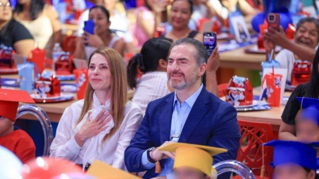 Man in a blue blazer and a woman in white sit in a crowded graduation ceremony others take photos in the background   Informe Regio