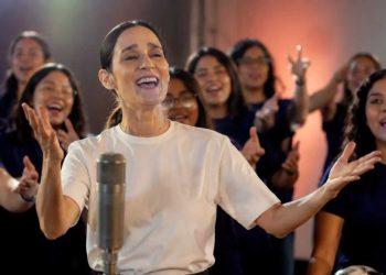Woman in a white shirt sings into a microphone with a cheering crowd behind her, hands raised and smiling families in the background.