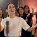 Woman in a white shirt sings into a microphone with a cheering crowd behind her hands raised and smiling families in the background   Informe Regio