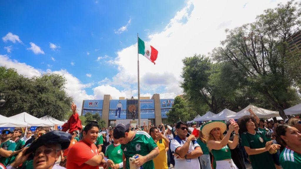 Crowd of people at an outdoor festival under a tall Mexican flag with white tents and a blue building in the background   Informe Regio