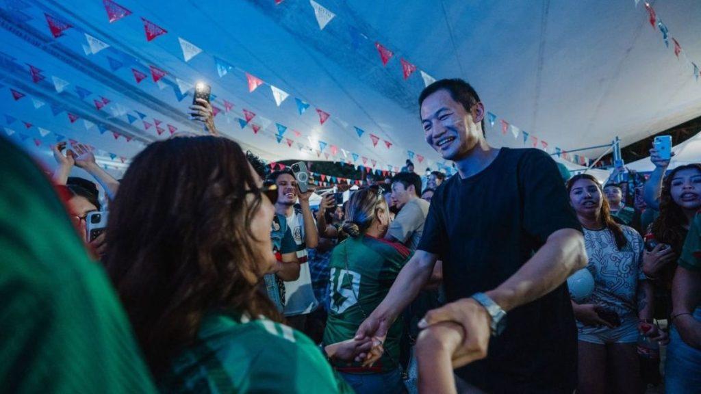 Man in a black shirt shakes hands with a crowd during a festive under tent celebration smiles around him as people take photos with their phones   Informe Regio
