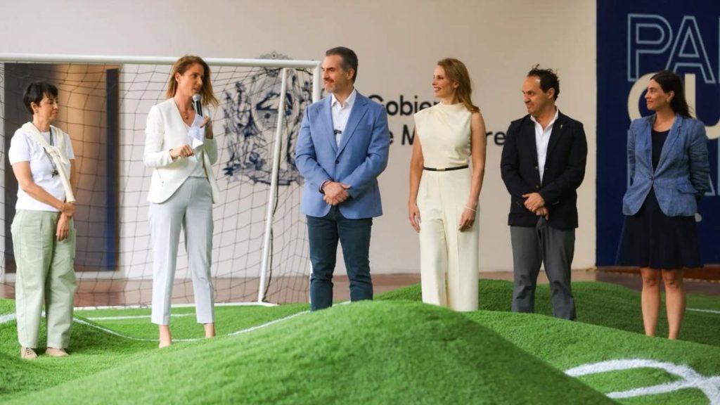 Speaker in a white blazer addresses five colleagues on an indoor artificial turf field a soccer goal behind them   Informe Regio