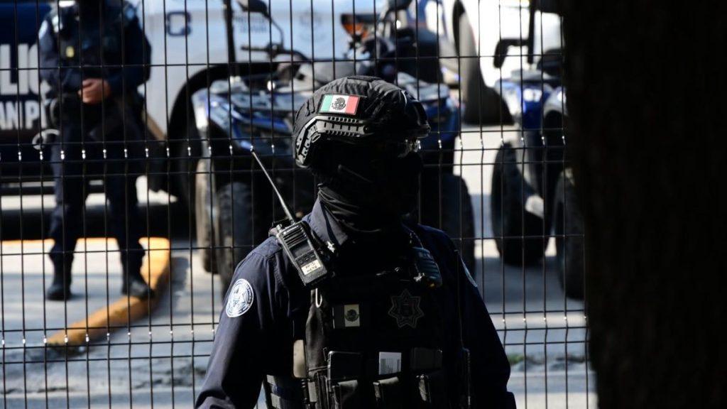 Tactical police officer in protective gear and helmet behind a metal fence with police vehicles in the background   Informe Regio